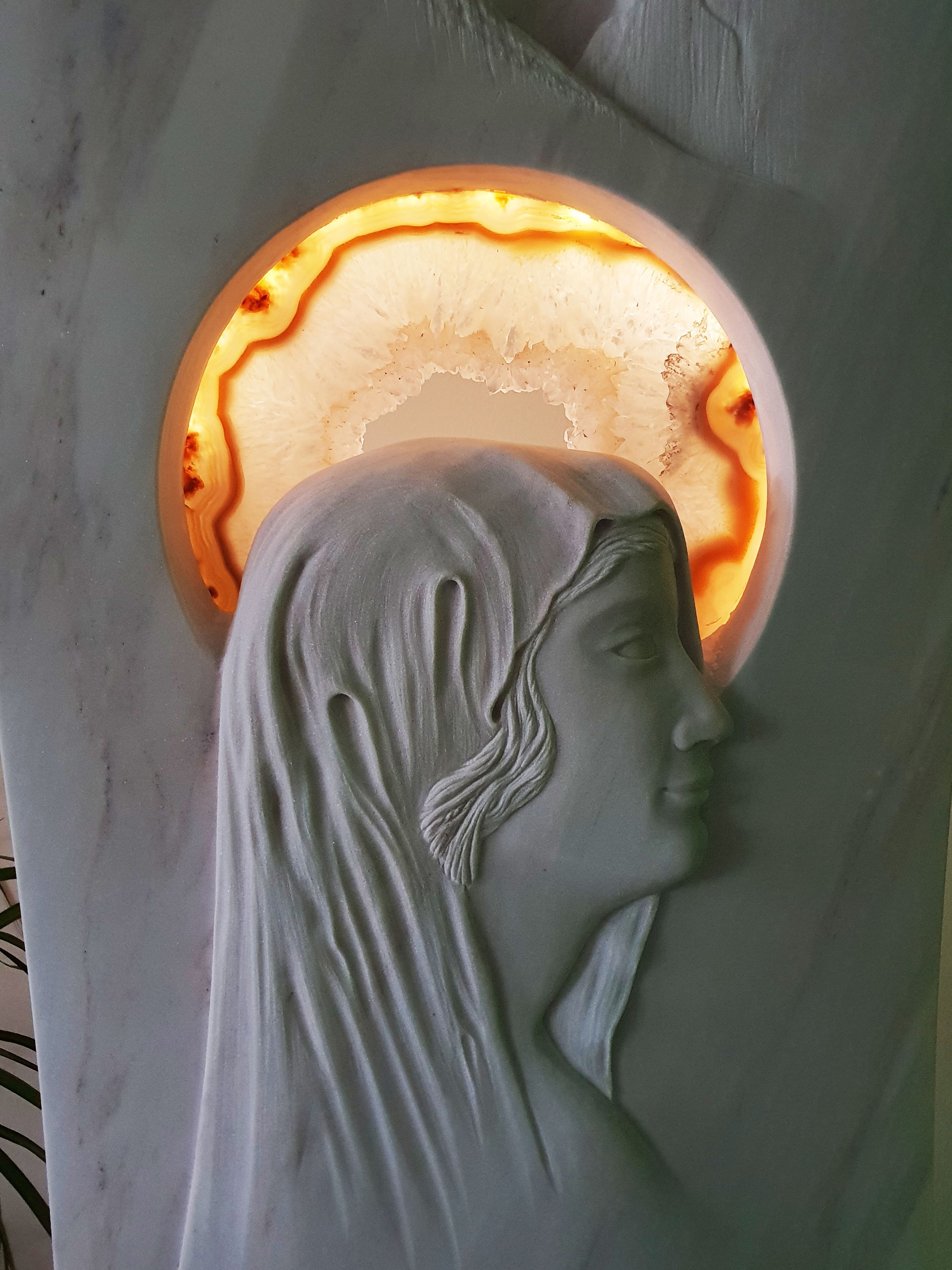 Einzigartige Grabmalkunst aus weissem Stein mit einer Heiligenfigur (Maria). Statt eines Heiligenscheins hat der Künstler einen anderen Stein in orangenen und roten Farbtönen in das Grabmal integriert und hinterleuchtet, handgefertigt von der Bildhauerei Sini in Grenchen, Detailansicht