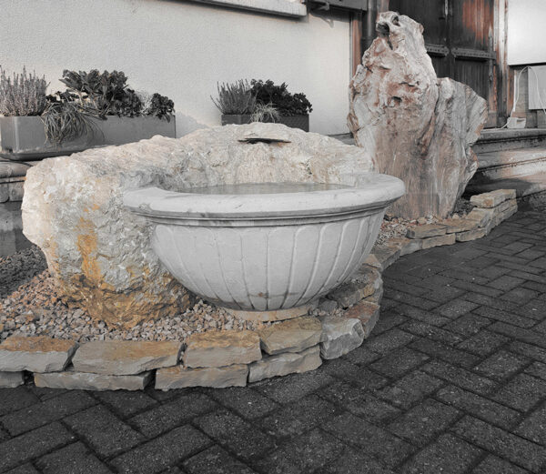 Ein dekorativer Brunnen aus Naturstein mit einer römisch inspirierten weissen Vase der vor einem traditionell ländlichen Haus steht, gestaltet von Bildhauerei Sini in Grenchen. (Region Solothurn und Bern)